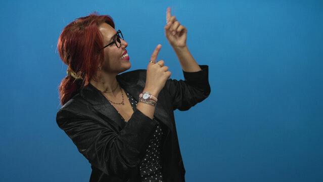 Young hispanic woman points finger toward upper right in blue studio while smiling, hands visible and wearing glasses with a black blazer; joy confidence.
