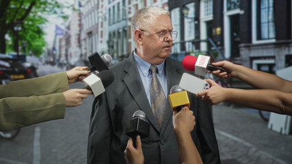 Naklejka premium Elderly man in suit surrounded by microphones and reporters' hands, raises hand while speaking on a street press interview; tension accountability.