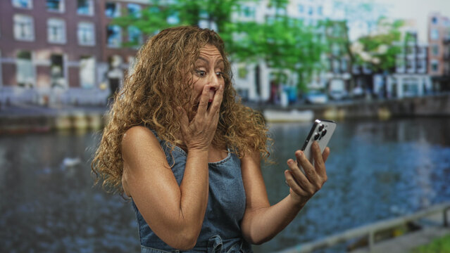 Woman holding smartphone, hand covering mouth in a shocked gesture by canal building with waterfront and row houses in view; surprise curiosity.