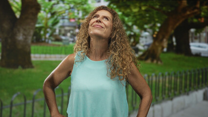 Woman middle age hispanic stands hands on hips looking up on a tree-lined street by a park fence wearing turquoise sleeveless top and curly hair  serenity. © Krakenimages.com
