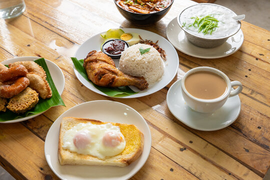 A vibrant spread of Malaysian favorites, Nasi Lemak Ayam Goreng, soft-boiled eggs on toast, assorted kuih muih, cendol, and a cup of teh tarik, served on a warm wooden tabletop.