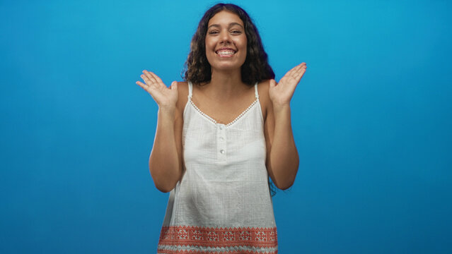 Woman clapping hands and smiling in a blue studio wearing white dress, hands near face and chest visible; joy surprise.