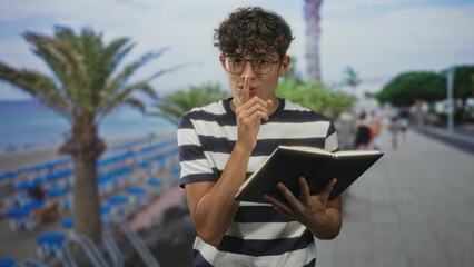 Fototapeta premium Teen boy in striped shirt and round glasses holds an open notebook and places finger to lips in a shushing gesture on a palm lined street by the beach; quiet mischief.