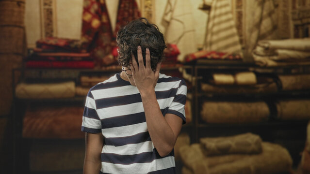 Teen boy touching face with hand inside a building carpet shop surrounded by rolled rugs and shelves; shy introspection.