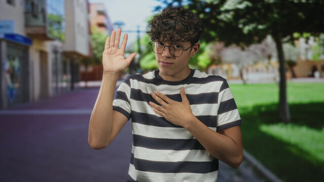 Teenage boy wearing glasses and a striped shirt places hand on chest and raises other hand on street; sincerity pledge.