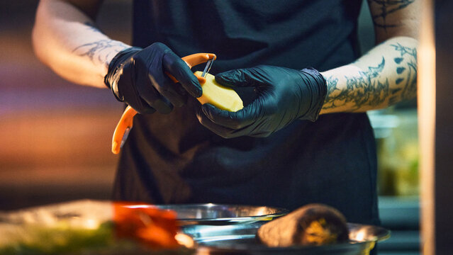 Chef peeling potato with orange peeler in moody restaurant kitchen. Concept of handmade food prep, catering mise en place, recipe production and culinary training content.