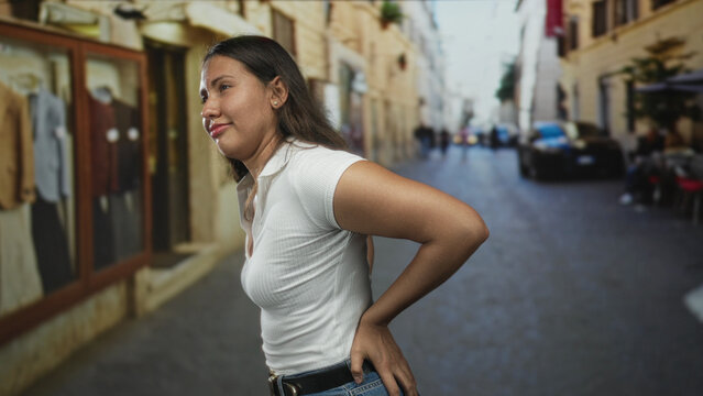 Woman holds lower back for backache on street storefront, side profile and hand on belt; discomfort routine.