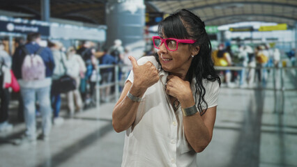 Obraz premium Woman with bright pink glasses and thumb pointing over shoulder in a busy airport queue, senior traveler in white blouse and bracelets; playful curiosity.