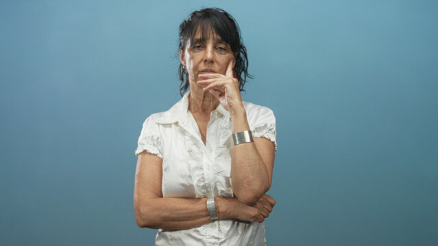 Woman senior hispanic with hand to mouth and crossed arms wearing white blouse and silver cuff in studio; contemplation resilience.