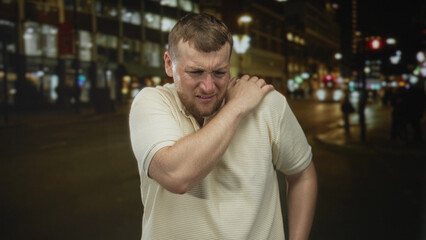 Man clutching his shoulder and grimacing on a busy city street at night under streetlights and blurred cars  pain. © Krakenimages.com