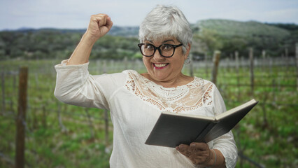 Naklejka premium Woman holding open book with raised fist, smiling in a vineyard with trellis posts and rolling hills, wearing glasses and lace blouse; joy.
