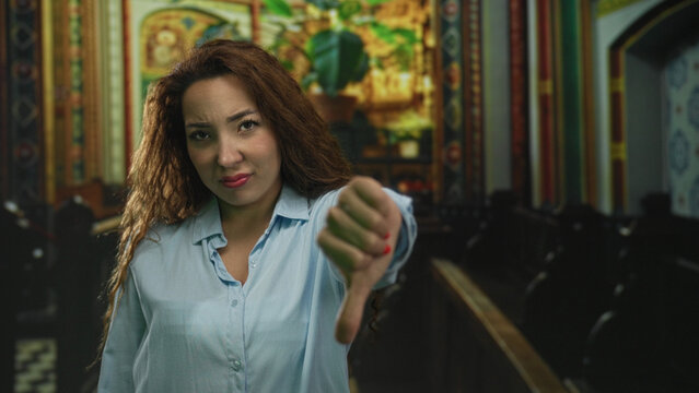 Woman giving thumbs down gesture toward camera in ornate church building nave with pews and painted iconography; disapproval protest.
