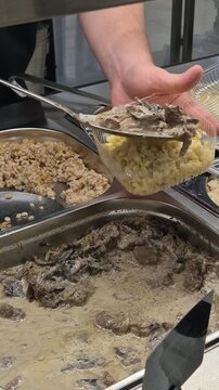 Buffet serving food worker placing creamy mushroom stew over pasta in a plastic take out container at a cafeteria
