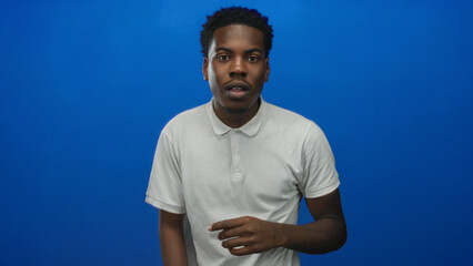 Fototapeta premium Young african american man in white polo shirt wiping eye gesture in vivid blue studio setting; sadness.