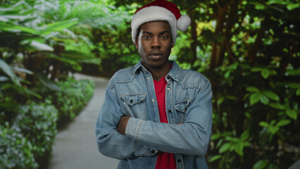 Naklejka premium Man wearing denim jacket and santa hat stands with crossed arms amid lush leafy greenery in park; holiday calm.