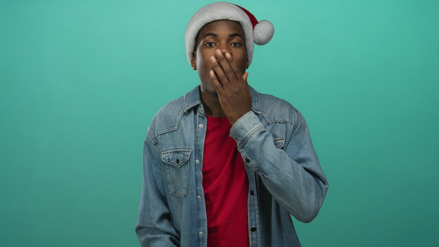 Young african american man in santa hat and denim jacket covering mouth with hand in teal studio; surprise.