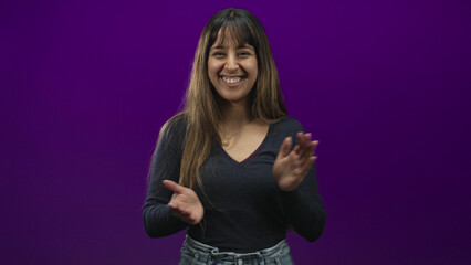 Naklejka premium Woman clapping hands in a purple studio setting, smiling with long hair, wearing v neck top, necklace and jeans in casual pose; joy celebration.