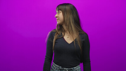 Naklejka premium Young woman wearing pendant necklace and high waisted jeans turns head to side looking left against a vibrant purple studio backdrop; contemplation.