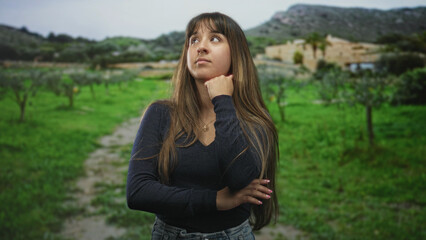 Obraz premium Woman with hand on chin and long hair covering face in front of building on a grassy path; quiet introspection.