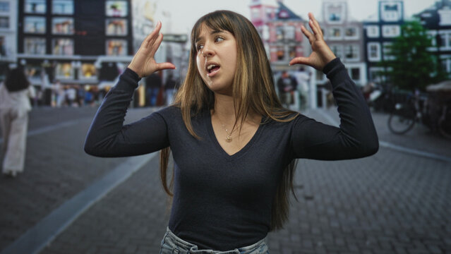 Young woman raises both hands beside her head, grimacing and showing a necklace on a cobbled street with canal houses; confusion uncertainty.