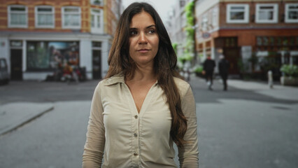Fototapeta premium Woman touching hair with right hand on a quiet street lined with brick shops and pedestrians, neutral expression and casual stance; thoughtful.