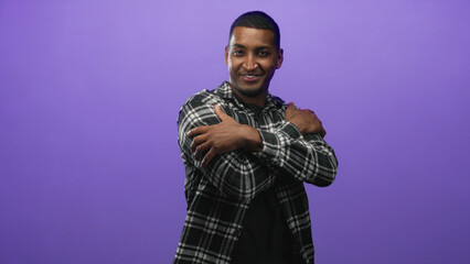 Fototapeta premium Young black man extends open hands and smiling face toward camera, plaid shirt visible as he reaches for an embrace in a purple studio; warmth connection.
