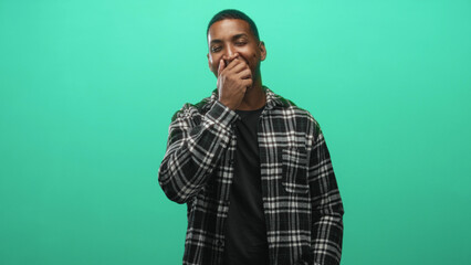 Fototapeta premium Man covering mouth with hand wearing a black plaid shirt against a green screen studio wall; playful amusement.