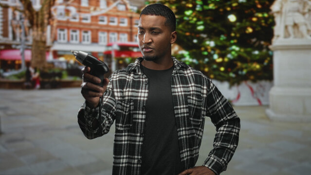 Man holding electric screwdriver with right hand, wearing plaid shirt, studying drill tip while hand rests on hip on street near a decorated christmas tree; concentration repair.