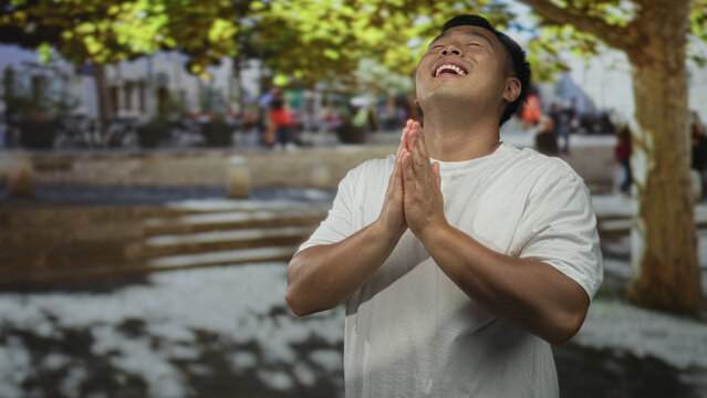 Man praying with clasped hands and closed eyes on busy city street by sidewalk and buildings under green canopy; gratitude.