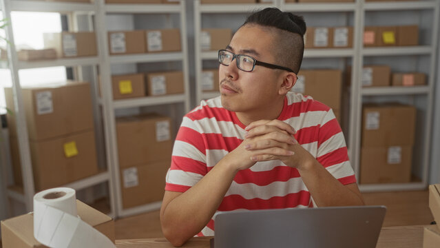 Man clasping hands over laptop and parcel at a desk with cardboard boxes on shelves in a building, wearing glasses and striped shirt; thoughtful planning.