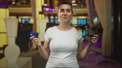 Woman in white t shirt holding a teal and black gamepad and blue creditcard with hands visible, smirking slightly in a studio set  playful consumerism. © Krakenimages.com
