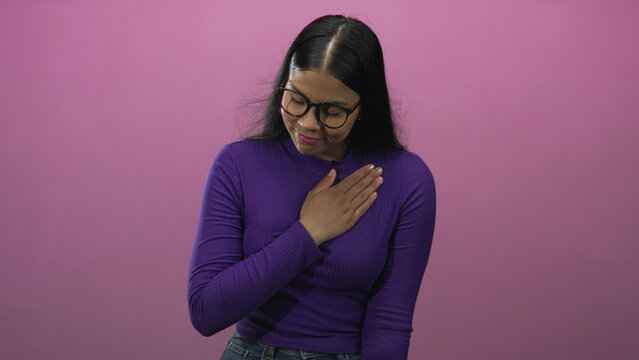Woman in purple sweater and glasses places hand on chest against smooth pink studio wall; sincerity pledge.