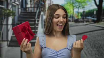 Naklejka premium Teenage girl holds gift box and heart in hands while smiling on busy urban street under sunlight; romance joy.