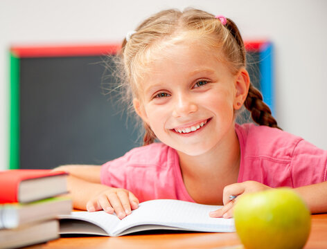 Little Schoolgirl Sitting At A Desk And Smiling Joyfully