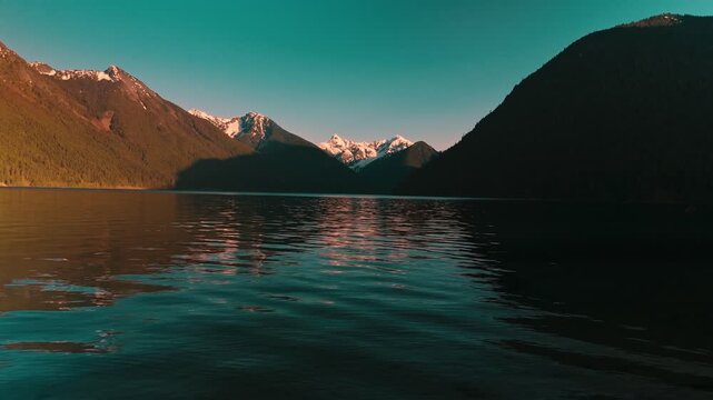 Deep dark pristine lake waters gently rippling extending toward spectacular rugged alpine mountains featuring bright white snow patches glowing from golden evening sunlight low altitude aerial flight