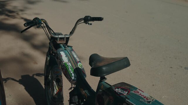 Worn and rusty moped with stickers on fuel tank and worn leather seat parked on sandy ground in Morocco