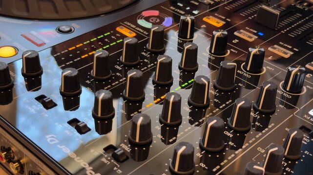Close-up view of a professional DJ mixer board showing multiple black knobs, sliders, and buttons illuminated by warm club lighting.