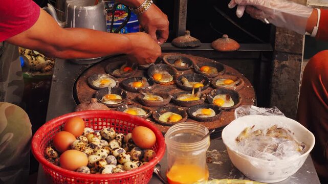 Traditional Vietnamese mini pancakes, known as Banh Can, being cooked on a clay stove. Vendors prepare the savory dish with eggs and shrimp at a local street food market