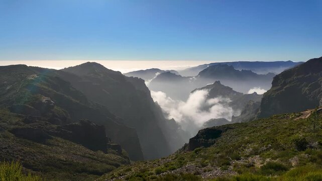 Aerial static shot captures a deep mountain valley on Madeira island, Portugal, with rugged cliffs dropping to a sea of clouds under a clear blue sky.