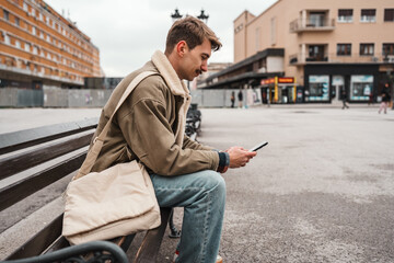 Obraz premium A young man with a mustache and a messenger bag sits on a wooden bench in a public urban square while focused on his mobile phone.