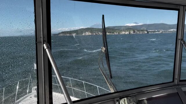 Point of view from a small boat cockpit with raindrops on the windshield, crossing choppy sea toward a rugged volcanic coastline in Kamchatka, Russia on a clear day