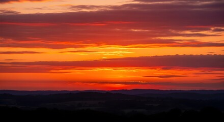 Fototapeta premium Vibrant orange and yellow sunset with clouds and a silhouette of mountains or hills in the foreground