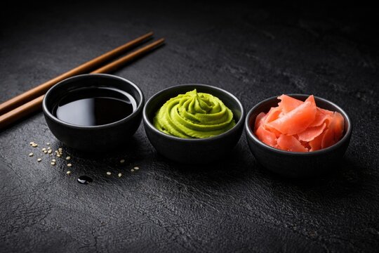 Set of soy sauce wasabi and marinated ginger bowls neatly aligned on a black textured backdrop with copy space