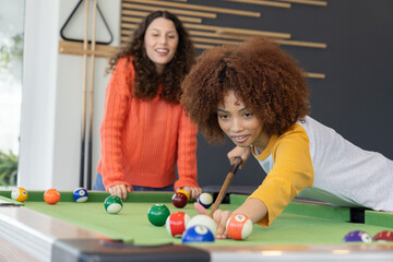 Naklejka premium Diverse female friends playing at rec room pool table with cue and balls, wearing colorful shirts