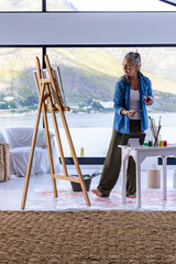 Naklejka premium Senior Asian woman wearing denim standing barefoot at easel in studio painting with brush and glass