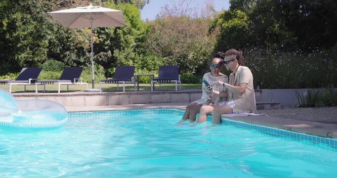 African American couple holding phone poolside in swimwear, swiping, toasting glasses, copy space
