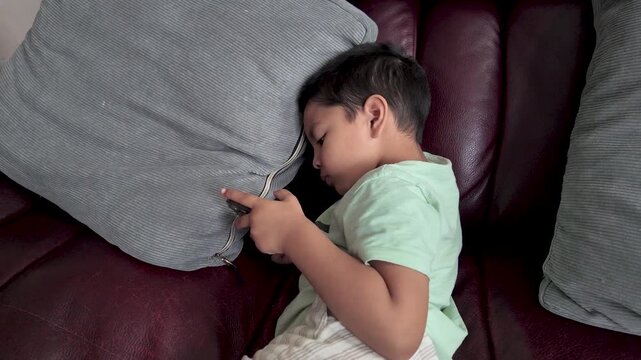 Child screen time and the impact of technology on early development. A young South Asian boy is engrossed in watching a smartphone while relaxing on a sofa at home.