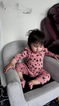 A cute South Asian baby girl is captured sitting on a small grey chair indoors, showcasing her early childhood development and innocent curiosity. She is dressed in pink pajamas with a leopard print.