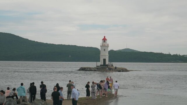 Static handheld shot of people walking along a narrow sandspit towards the Tokarevsky lighthouse in Vladivostok. Overcast maritime landscape with gentle waves and green hills in the background.