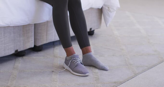 African American woman sitting on bed slipping foot into gray sneaker tying laces preparing workout